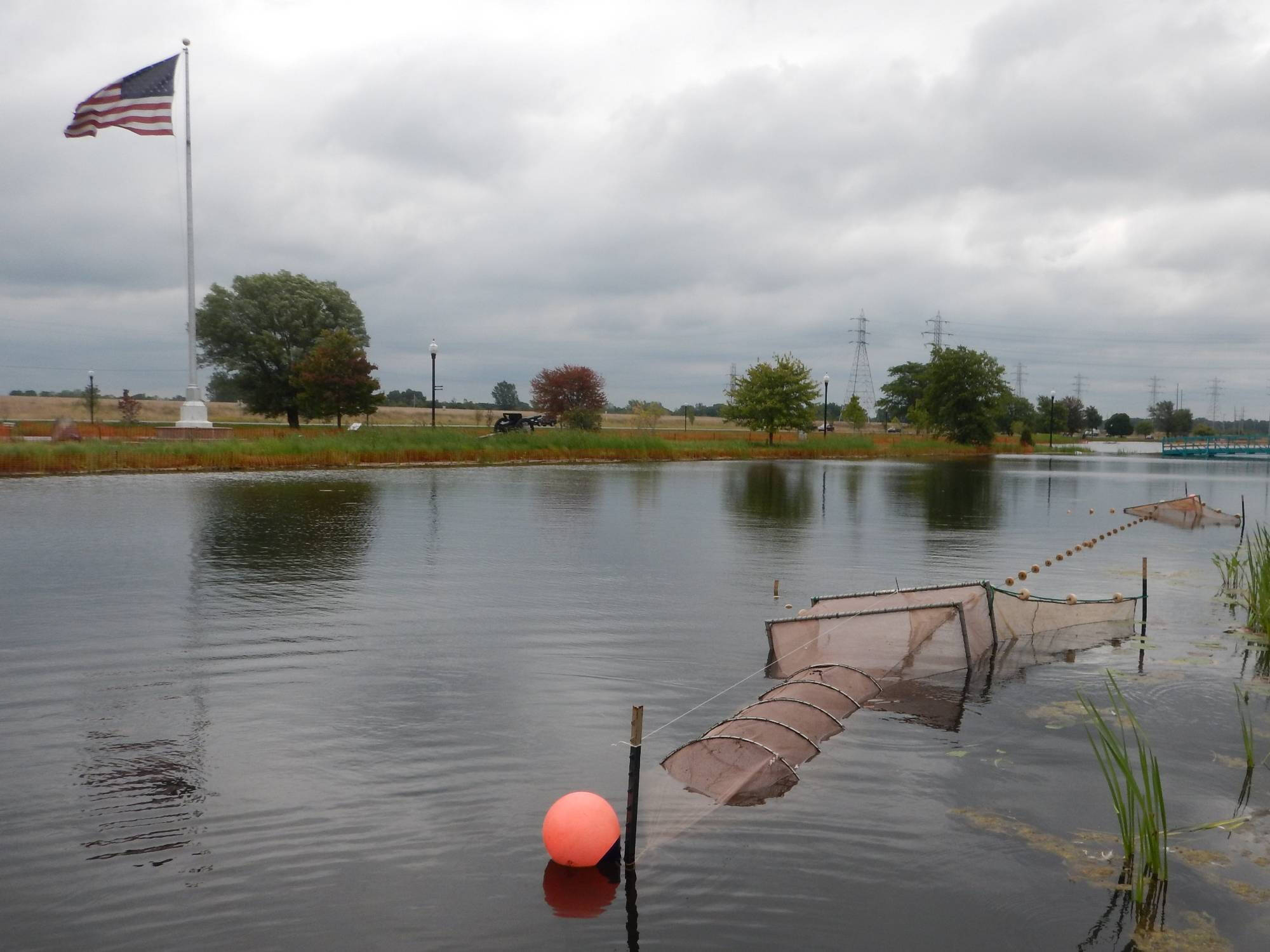 A fyke net is shown set up in an open water pond; an American flag and memorial cannon are seen in the distance.